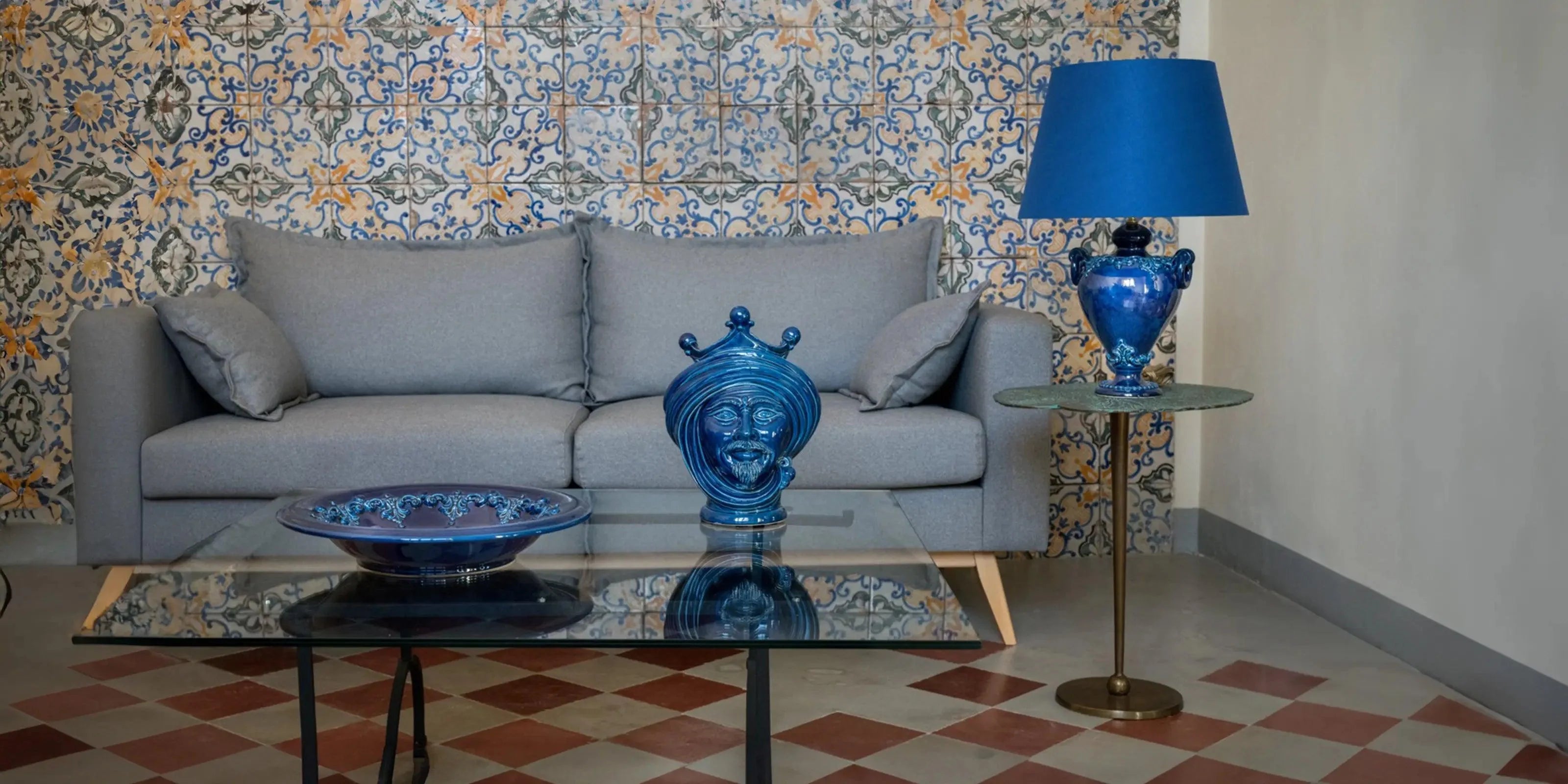 Soggiorno con divano grigio, piastrelle ceramiche Caltagirone fatte a mano, e oggetti blu su tavolo vetro e lampada